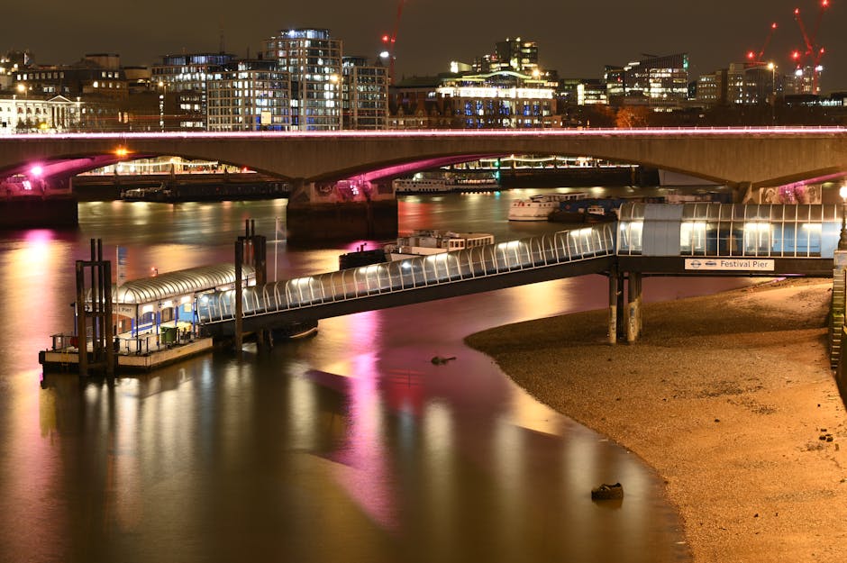 Nighttime view of the Festival Pier on the Thames River near Waterloo Station SE1, featuring a modern glass-covered riverside walkway extending over the water, illuminated with bright white lights. Adjacent to the walkway is a sandy riverbank with a few scattered rocks, while the calm water reflects the vibrant pink and yellow lights from nearby buildings. In the background, a large bridge with pink accent lighting spans across the river, and the city skyline is dotted with multi-story buildings, some under construction with cranes visible. The scene showcases a combination of urban infrastructure and water-based leisure spaces, highlighting the area's vibrant night-time atmosphere.