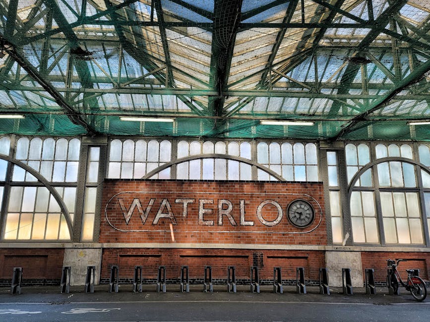 Exterior view of Waterloo Station with a large brick wall featuring the illuminated 'Waterloo' sign and a clock. The station's glass and metal roof structure, with visible green framing, covers the area. The space appears clean and well-maintained, with bicycle parking racks and a bicycle visible on the right side of the image. The lighting is natural, and the overall atmosphere conveys a tidy, hygienic transportation hub, reflecting proper surface cleaning and maintenance by Waterloo Carpet Cleaning. This image highlights the station’s architectural details and cleanliness, suitable for content related to professional cleaning and hygiene standards near Waterloo Station SE1, WATERLOO.