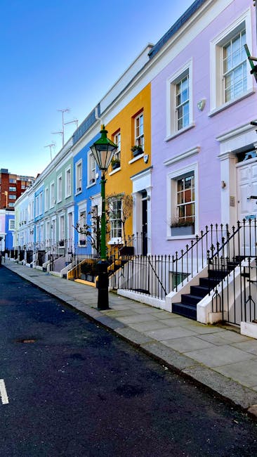 A row of colorful Victorian-style terraced houses along Lower Marsh in Waterloo, with pastel facades in shades of purple, yellow, and light blue. Each house features white window frames and decorative cornices, with small front gardens bordered by black wrought iron railings. A traditional black lamppost stands on the pavement, and the street appears clean and well-maintained under clear blue skies. This residential area exemplifies the charming architecture in Waterloo, suitable for professional domestic cleaning and surface sanitisation as provided by Waterloo Carpet Cleaning.