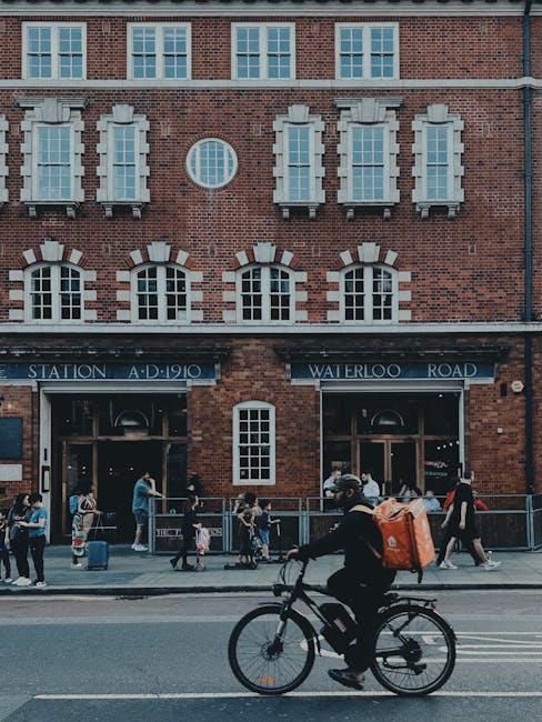 Photograph of a brick building with multiple white-windowed stories and a circular window. The ground level features a station entrance labeled 'Waterloo' with a dark awning displaying 'STATION A.D. 1910' and 'WATERLOO ROAD'. Several pedestrians are visible on the sidewalk, including a cyclist wearing a helmet and carrying a large orange bag, riding past the station. The scene is illuminated by natural daylight, and the overall setting is urban and lively, showcasing a typical busy street near Waterloo station. This image relates to the domain waterloocarpetcleaning.co.uk and is relevant to the service category of cleaning, highlighting the importance of maintaining cleanliness in busy public and commercial spaces.