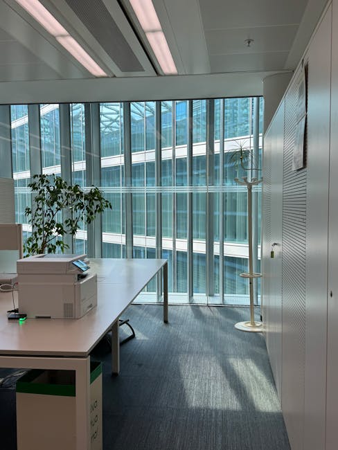 An office interior featuring a large floor-to-ceiling glass window with vertical blinds, allowing natural light to illuminate the space. In the foreground, a white desk holds a multifunction printer and a small green object, possibly a desk accessory. Next to the desk, a potted plant adds greenery to the room. The flooring is covered with grey carpet, appearing clean and well-maintained. A light ceiling with built-in lighting panels provides additional illumination. On the right side, there are white storage cabinets with horizontal slats, and a coat stand with a single hook. The overall environment reflects a modern, tidy, and professional office space with a focus on hygiene and surface cleanliness, which Waterloo Carpet Cleaning is equipped to support through their professional cleaning services in the Southbank Centre Waterloo area.