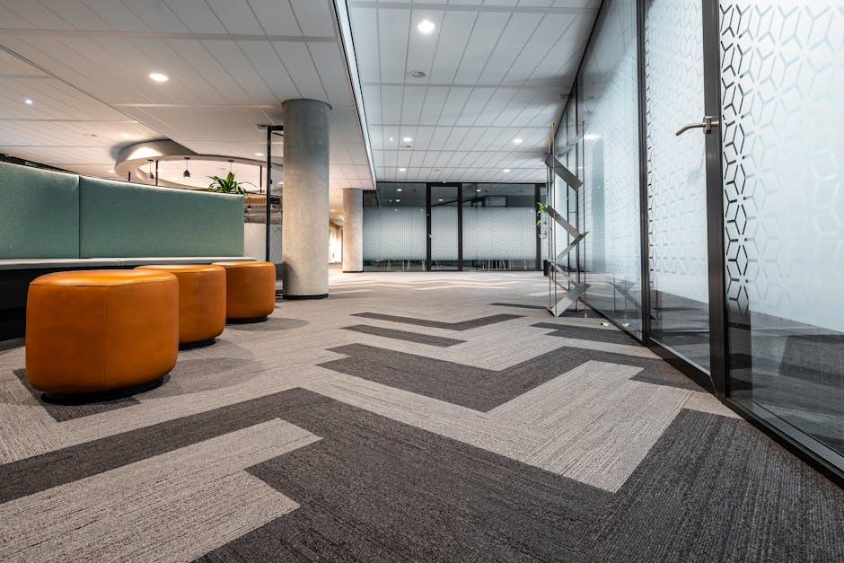 The image depicts a modern office lobby area with a patterned carpet flooring in shades of gray and beige, featuring geometric lines. On the left, there are three rounded, orange leather stools positioned near a curved, upholstered light teal seating area. Behind the seating, a large concrete column supports the ceiling, which has sleek, installed recessed lighting. To the right, a glass wall with a sliding door separates the lobby from an adjacent room, showcasing a decorative honeycomb pattern on the glass. The space appears clean, well-maintained, and well-lit, with a contemporary minimalist design. This setting exemplifies professional surface cleaning and maintenance, aligning with services offered by Waterloo Carpet Cleaning, as referenced on the page about office carpet cleaning at Southbank Centre Waterloo.