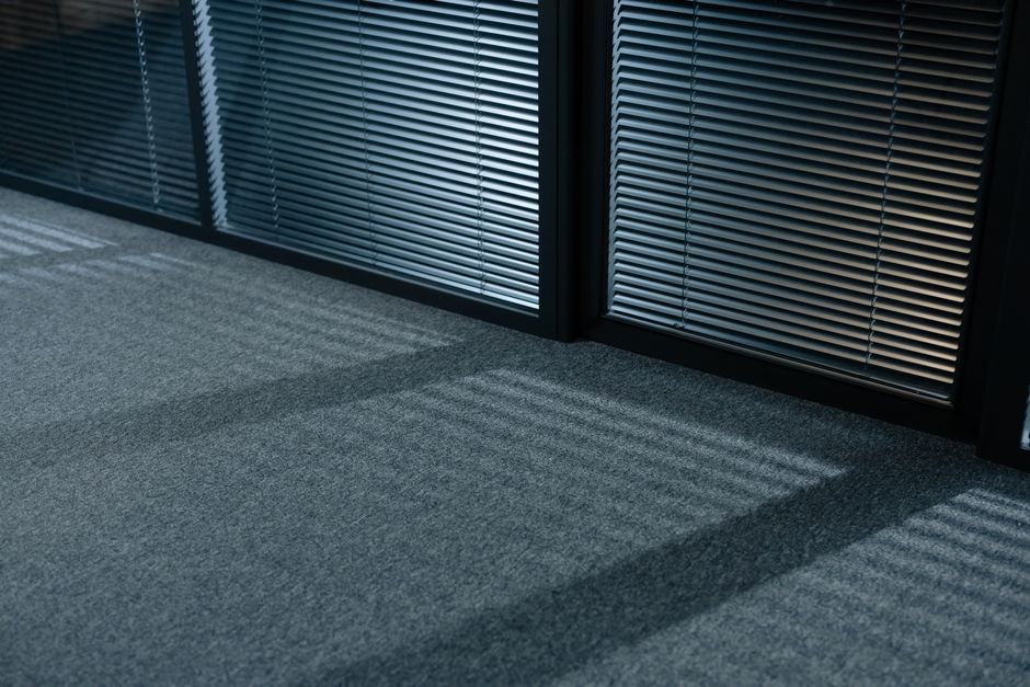 An office interior featuring a large floor-to-ceiling glass window with vertical blinds, allowing natural light to illuminate the space. In the foreground, a white desk holds a multifunction printer and a small green object, possibly a desk accessory. Next to the desk, a potted plant adds greenery to the room. The flooring is covered with grey carpet, appearing clean and well-maintained. A light ceiling with built-in lighting panels provides additional illumination. On the right side, there are white storage cabinets with horizontal slats, and a coat stand with a single hook. The overall environment reflects a modern, tidy, and professional office space with a focus on hygiene and surface cleanliness, which Waterloo Carpet Cleaning is equipped to support through their professional cleaning services in the Southbank Centre Waterloo area.