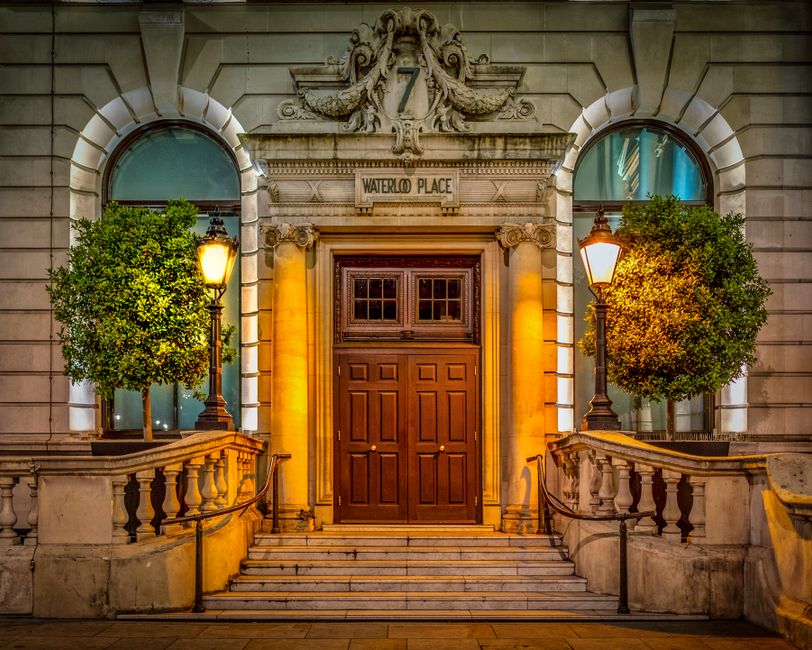 Image of the entrance to Waterloo Place, a historic building with ornate stone architectural details, including decorative carvings above the wooden double doors and a stone plaque reading 'Waterloo Place' situated between two arched windows. The entrance is accessed via a short flight of marble steps with metal handrails on each side. Two traditional-style lamp posts with warm yellow lighting illuminate the doorway, highlighting the well-maintained stone balustrades and lush green potted trees flanking the entrance. The scene is captured during evening hours, with soft ambient lighting enhancing the elegant and clean façade, exemplifying a tidy and well-kept residential or commercial property in Waterloo, London. Waterloo Carpet Cleaning provides surface cleaning and deep cleaning services for properties like this, ensuring hygiene and pristine conditions.